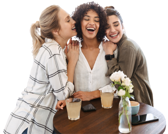 Three women smiling and embracing one another while standing at a table with drinks
